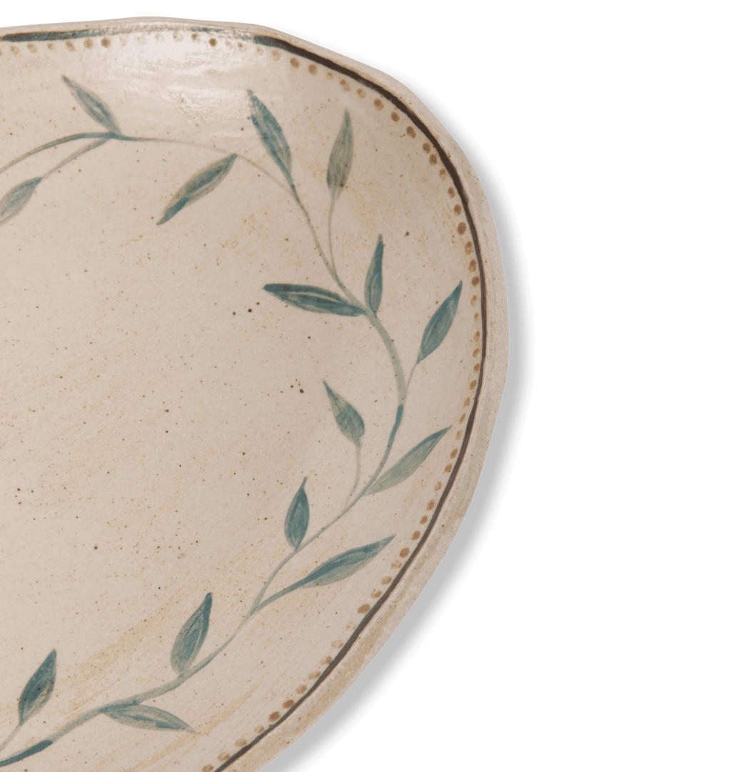 Close up detailed view of an oval ceramic serving platter with an organic rim, decorated with a green leafy vine border and a gold dotted accent line on a white background