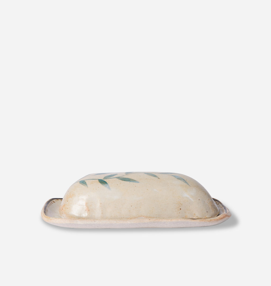 A small ceramic butter dish with a domed lid decorated with green leaf motifs, set against a plain light gray background.