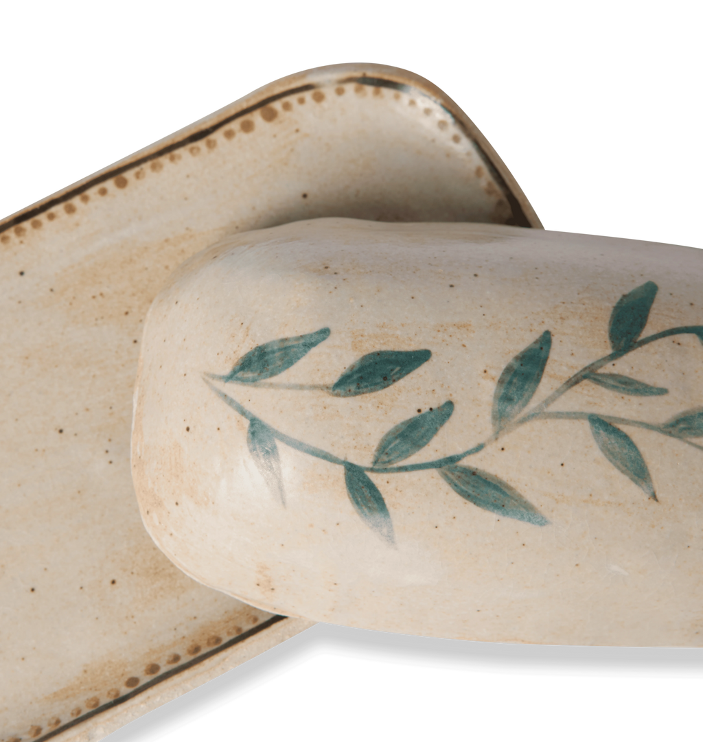 Close up view of a ceramic butter dish and lid decorated with a green leafy vine border and a gold dotted accent line on a white background