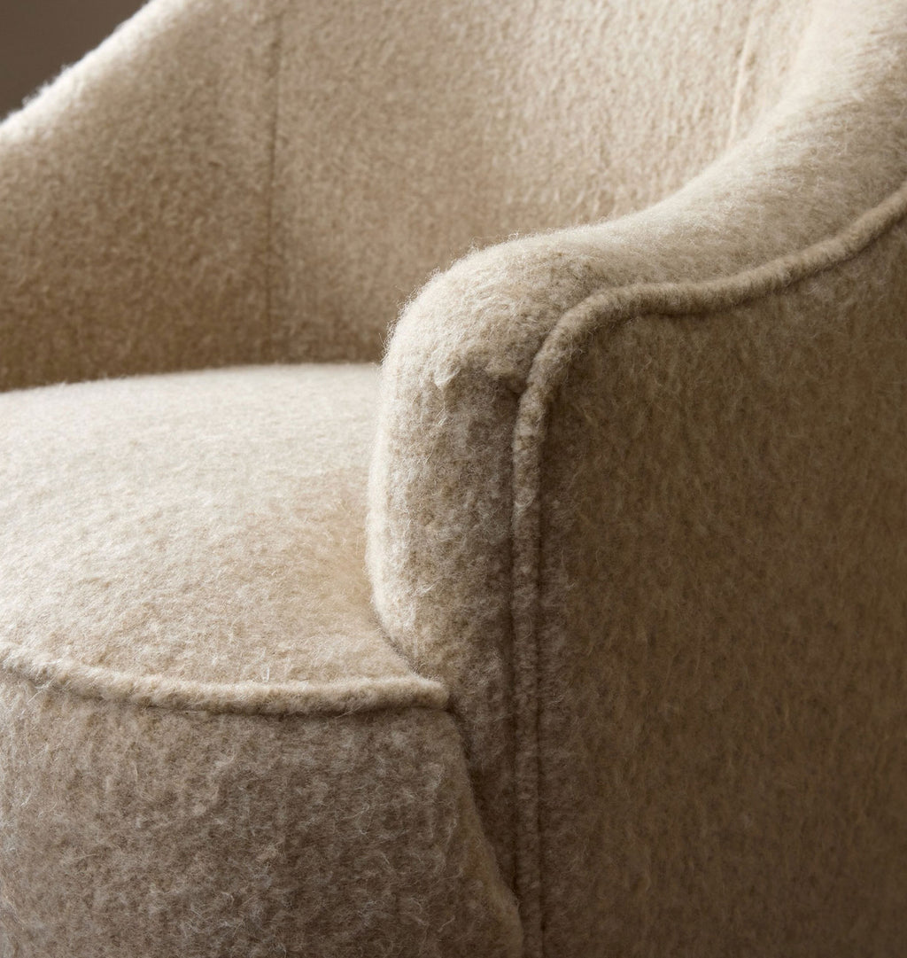 Close-up of a plush upholstered armchair in a warm beige or oatmeal-colored fabric, showing the soft texture and rounded arm detail.