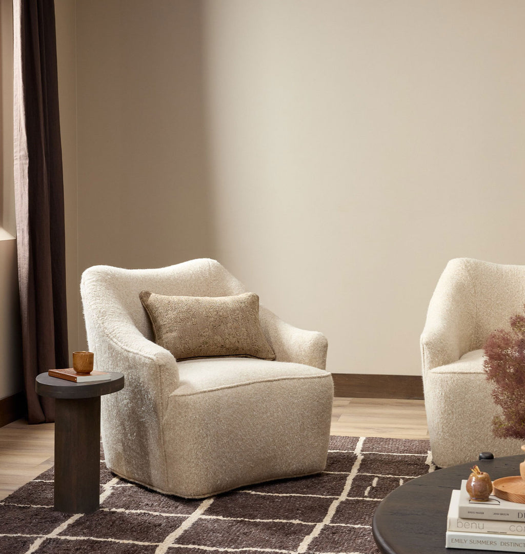 A plush upholstered armchair in a warm cream bouclé fabric with a matching throw pillow, styled in a minimalist living room setting with a dark wood side table and a patterned rug.