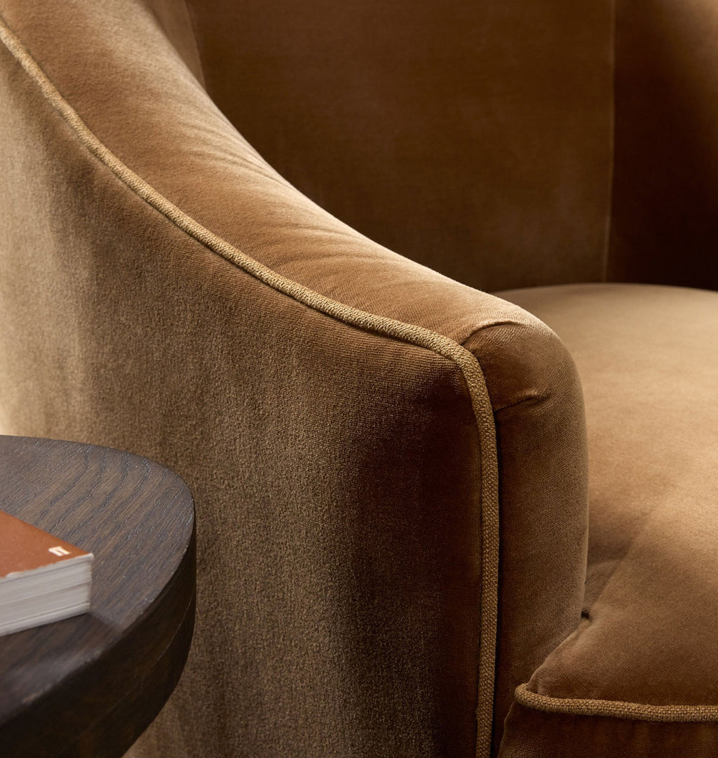 Close-up of a plush upholstered armchair in warm camel brown velvet, showing the curved arm, channel tufting, and side seam detail. A small dark wood side table with a book is partially visible in the lower left corner.