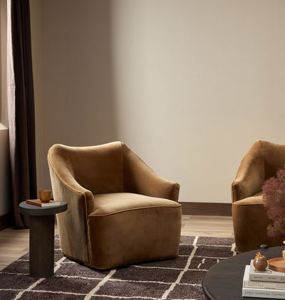 A modern curved armchair upholstered in warm camel velvet, styled in a minimalist living room setting with a dark side table, a patterned rug, and another matching chair partially visible.