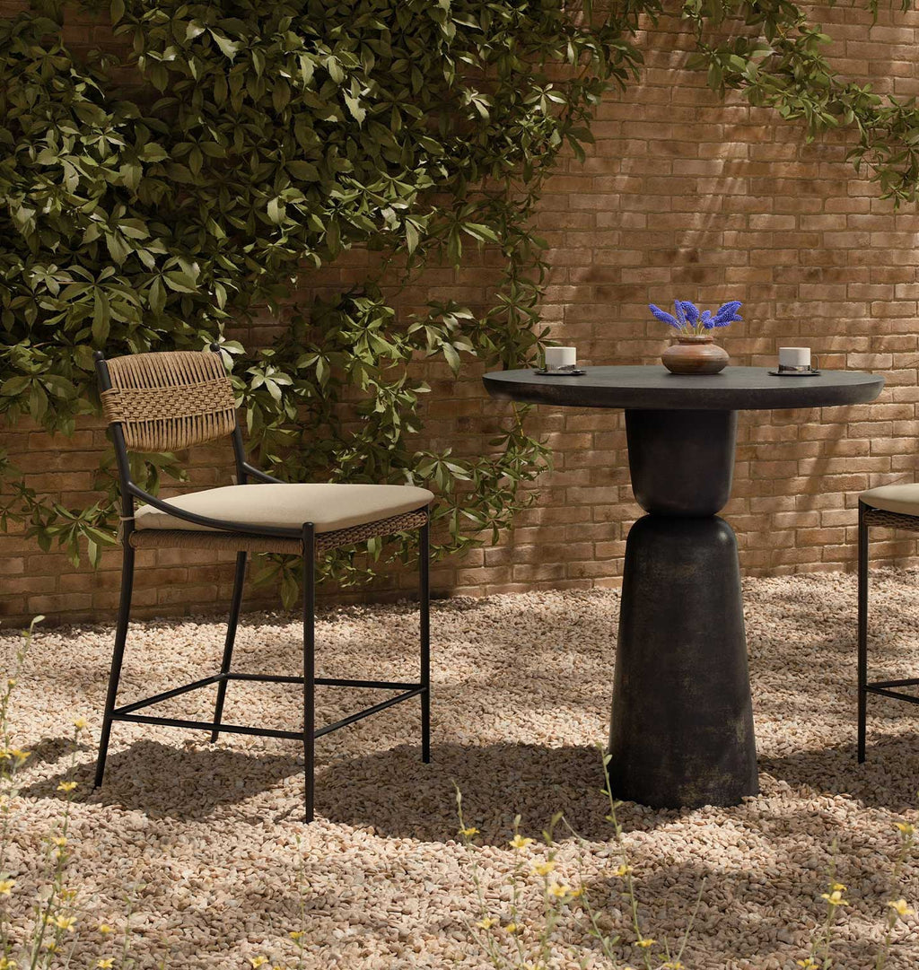 Outdoor patio furniture set featuring a tall barstool with a woven backrest and beige cushion, next to a round dark stone-top bar table with a pedestal base, set on a gravel surface against a brick wall and leafy greenery.