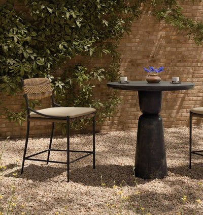 Outdoor patio furniture set featuring a tall barstool with a woven backrest and beige cushion, next to a round dark stone-top bar table with a pedestal base, set on a gravel surface against a brick wall and leafy greenery.