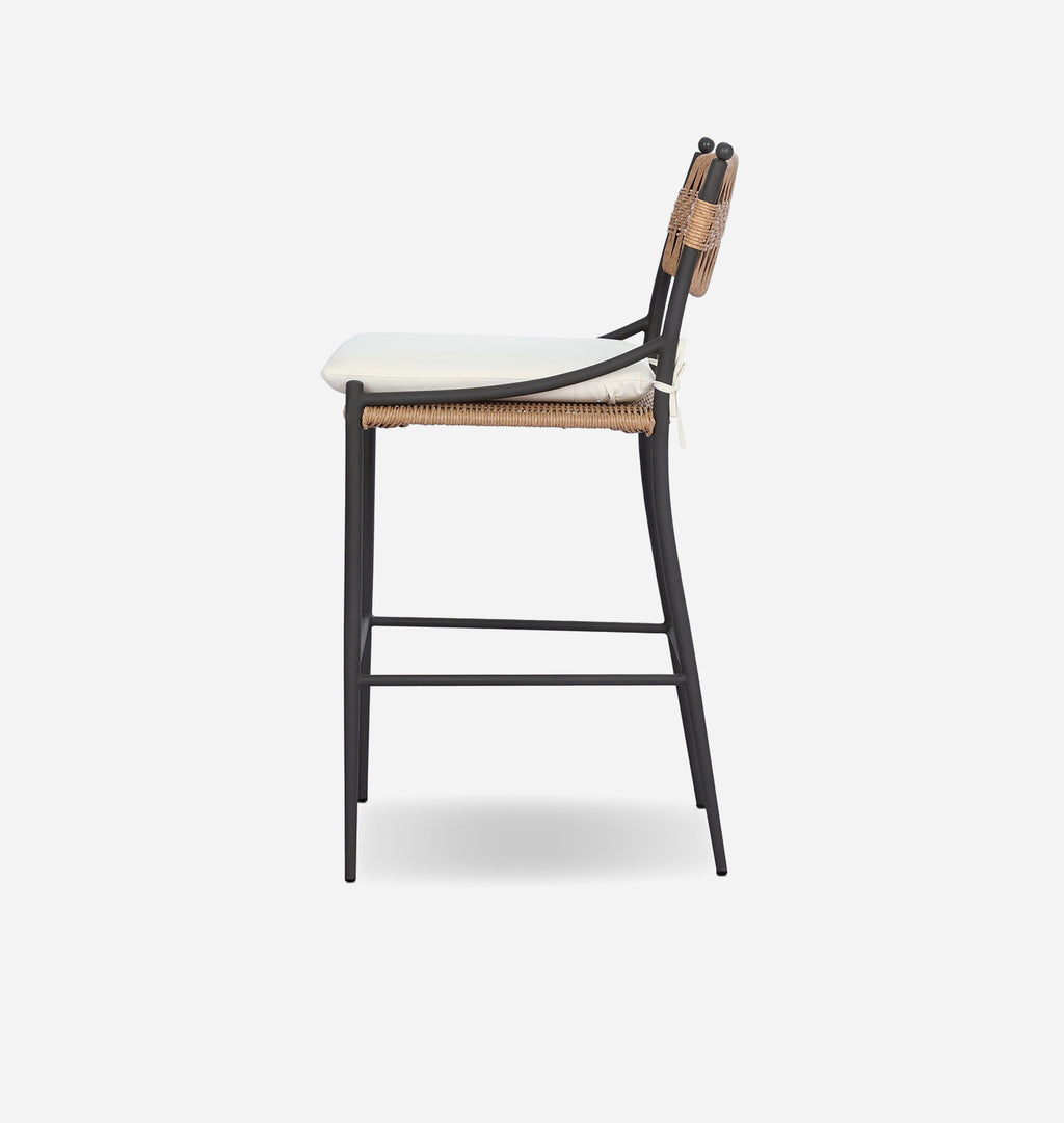 A modern counter height bar stool shown from a side profile view, featuring a black metal frame, woven natural seat and backrest, and a white upholstered seat cushion, set against a solid light gray background.