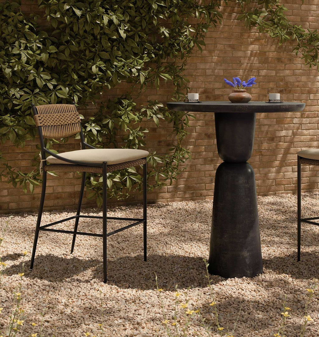 Outdoor patio furniture set featuring a round dark stone bar-height table and two matching bar stools with woven backs and beige seat cushions, set on gravel in front of a brick wall and leafy greenery.