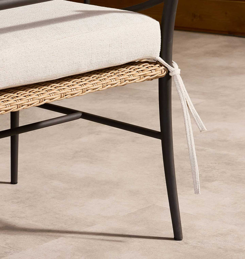 Close-up of a woven rattan chair seat with a light beige cushion tied at the side, set on a neutral beige floor.