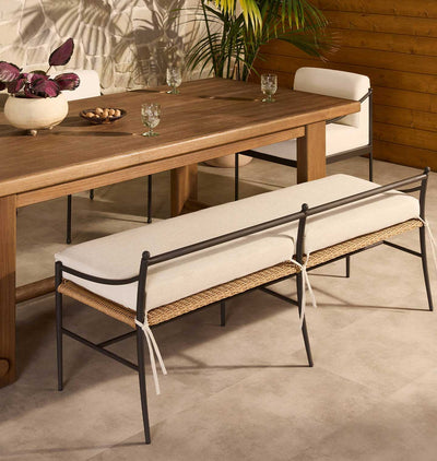 A modern outdoor dining bench with cream cushions and a woven rattan seat, positioned next to a wooden dining table in a patio dining room setting.
