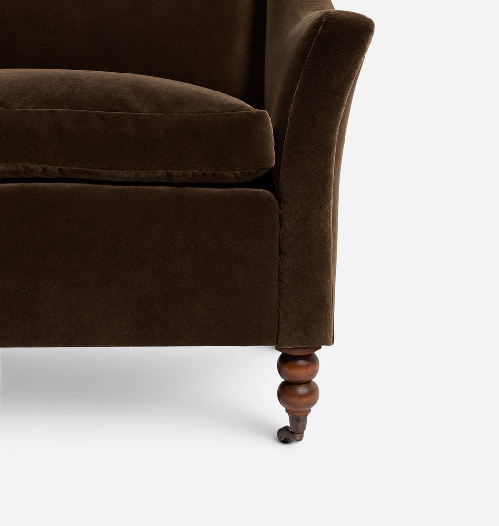 Close-up of a dark brown velvet armchair showing the corner of the seat cushion, rolled arm, and turned wooden leg on a light gray background.