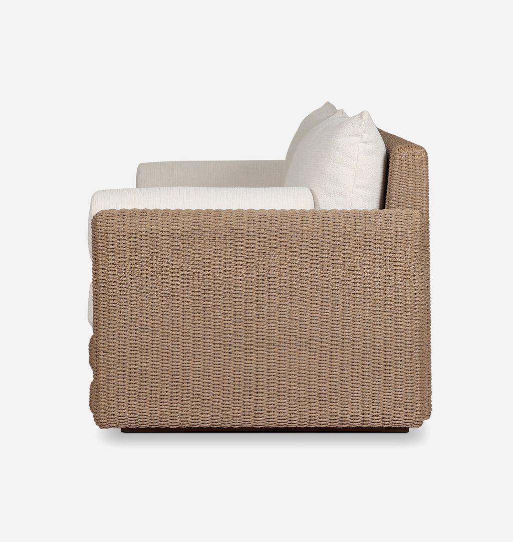 A modern outdoor loveseat with a woven wicker frame in a warm taupe-brown color, featuring thick off-white seat and back cushions, shown from a side angle against a plain white background.