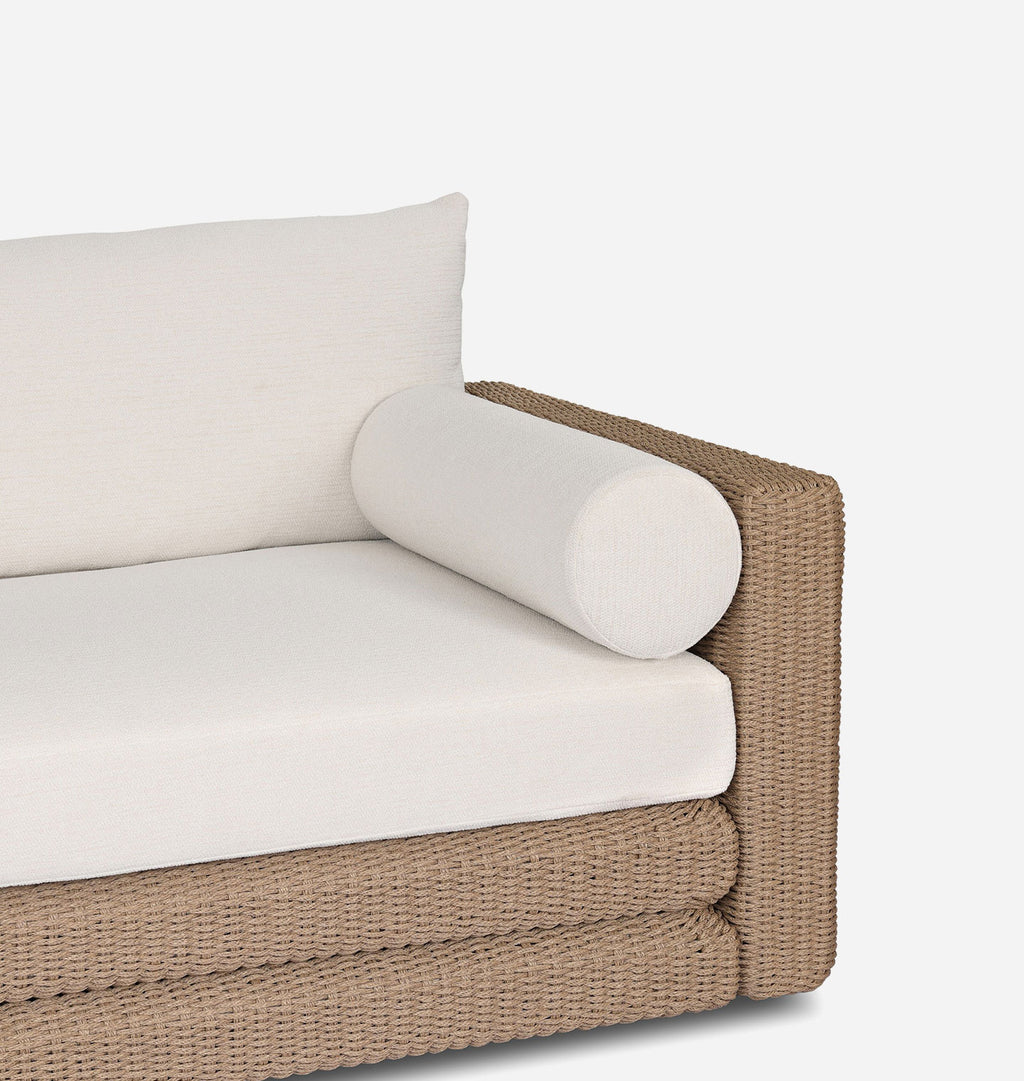 Close-up of a woven outdoor sofa corner with off-white seat and back cushions, a cylindrical back cushion, and a tan wicker frame on a light gray background.