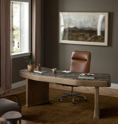 A curved wooden desk with a dark stone-look top and medium brown legs, styled in a home office with a brown leather swivel chair, a potted plant, and a framed landscape painting on a dark gray wall.