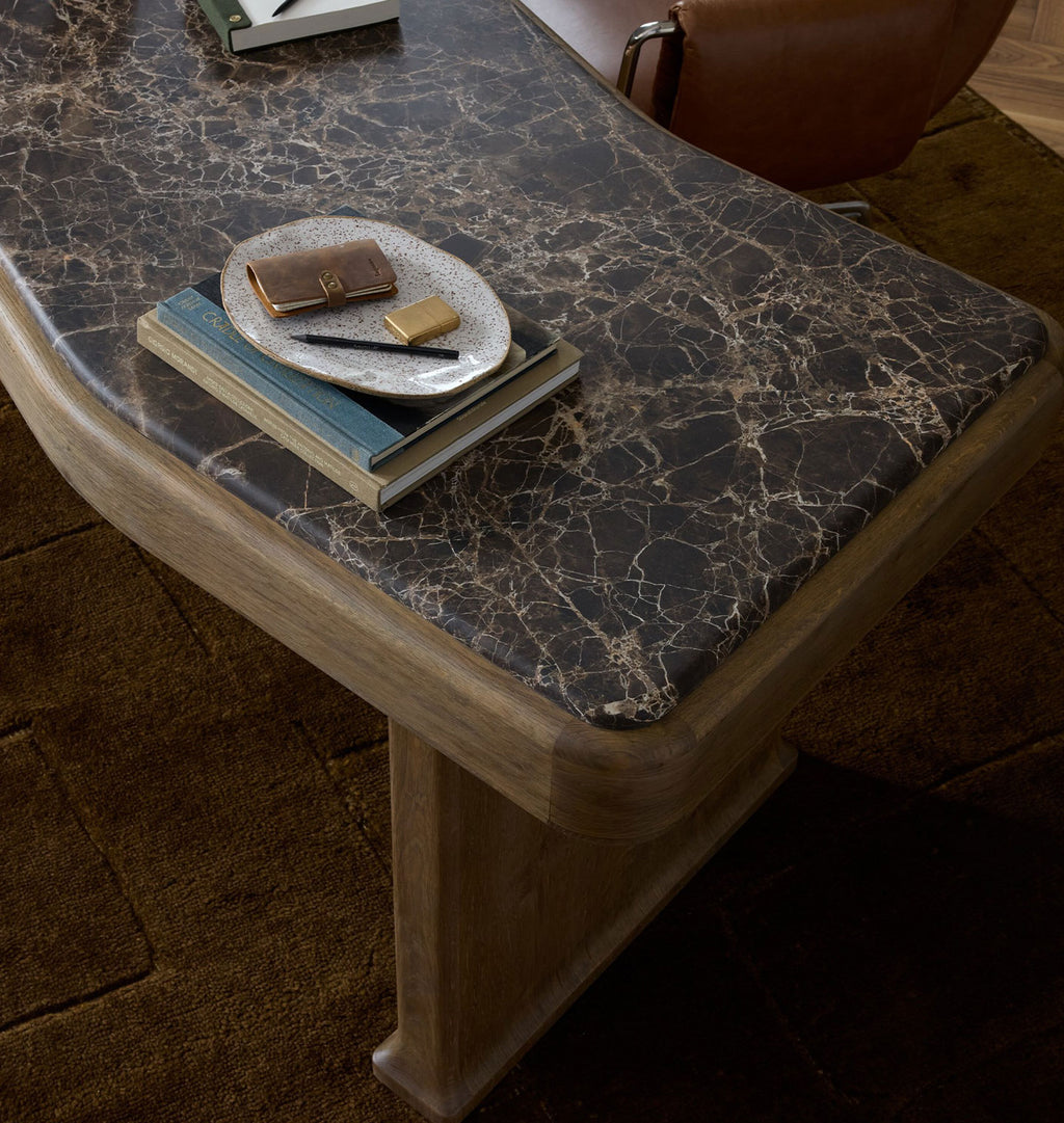 A close-up of a modern desk with a dark gray and black marble top and a light natural wood frame, styled with books and a small plate in a warm interior setting.