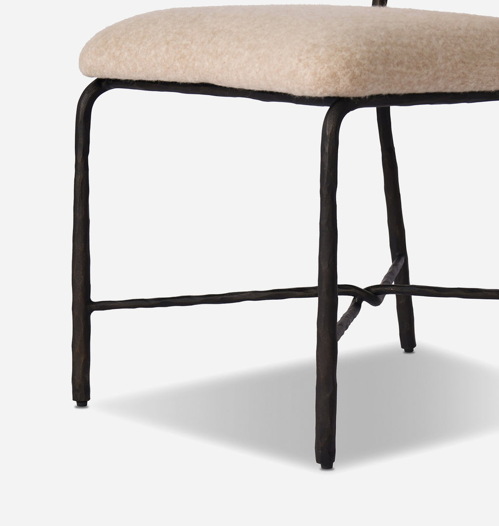 A close-up of a stool with a textured cream upholstered seat and a dark, rustic metal frame with a twisted leg design and horizontal stretcher bar.