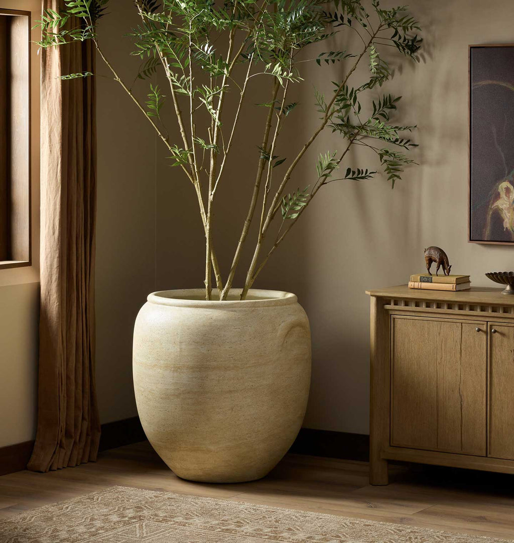 A large beige stone planter holding a tall bamboo or palm-like plant with slender green leaves, placed in a styled interior room with wood flooring, a curtain, and a wooden cabinet.