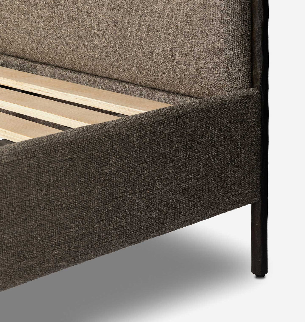 Close-up of a bed frame upholstered in textured taupe-brown fabric, showing the headboard, side rail, slatted wooden base, and dark metal leg against a light gray background.