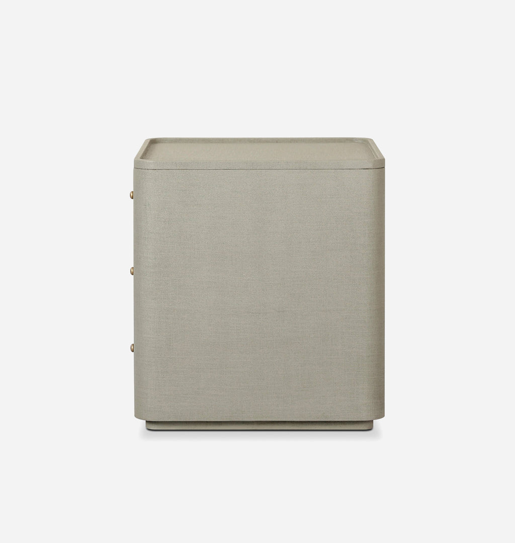 A modern, minimalist side table or nightstand with a rectangular top and three drawers, upholstered in a textured light beige fabric with small gold knobs, shown against a plain light gray background.