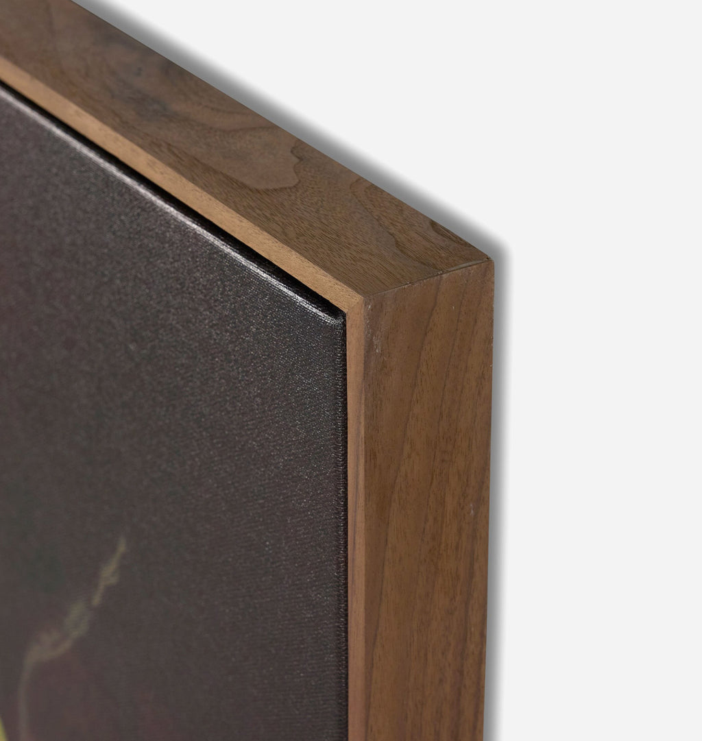 Close-up of a framed wall art piece showing the corner where a medium brown wooden frame meets a dark textured canvas or print, against a light gray background.