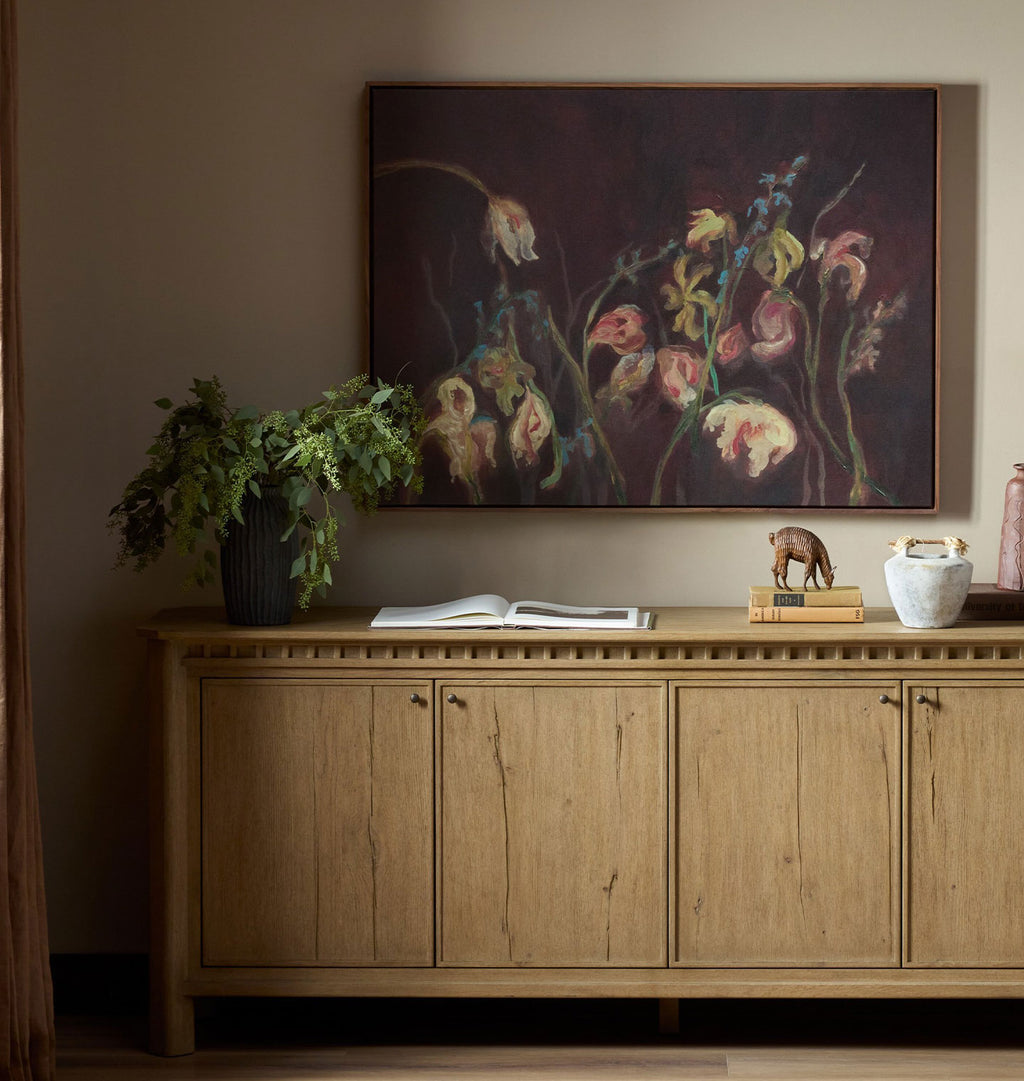 A framed floral painting hangs on a beige wall above a light wood sideboard. The artwork shows dark, moody flowers in pink, cream, and muted yellow tones against a deep brown background.
