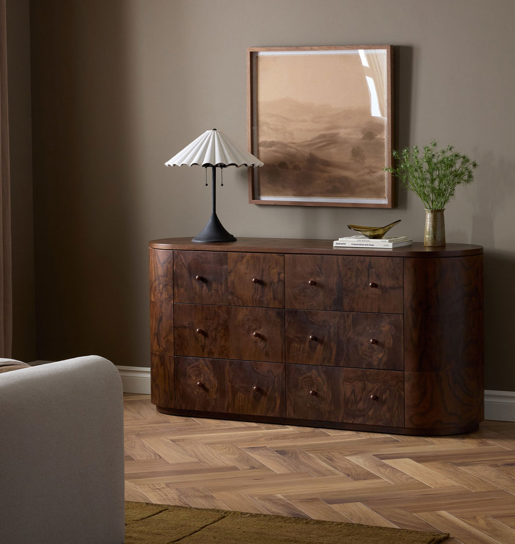 A curved dark wood dresser with six drawers and small round knobs, styled in a bedroom setting with a white pleated table lamp, a framed landscape print, books, and a vase of greenery.