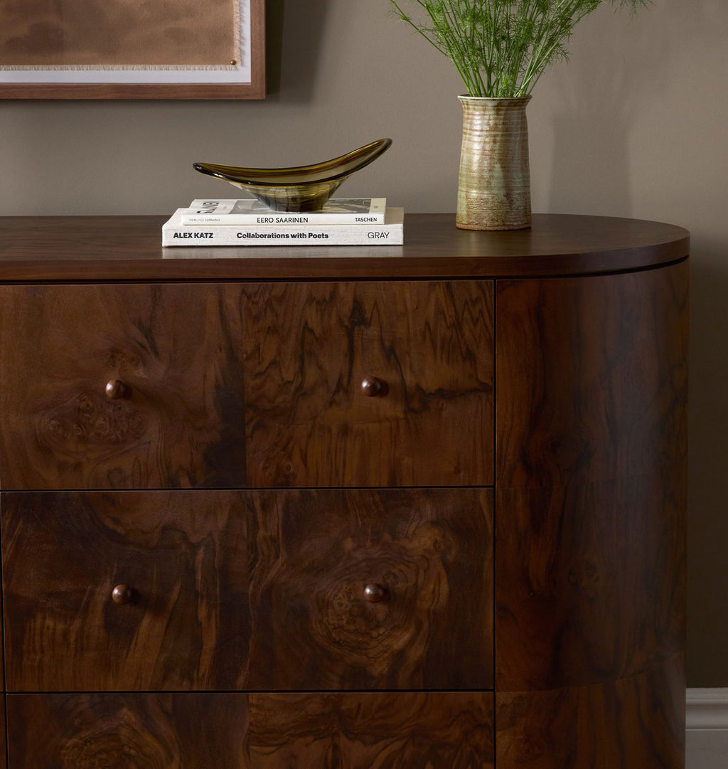 A curved wooden dresser with a rich dark brown wood grain finish, featuring three drawers with round knobs and styled with books, a bowl, and a vase on top.