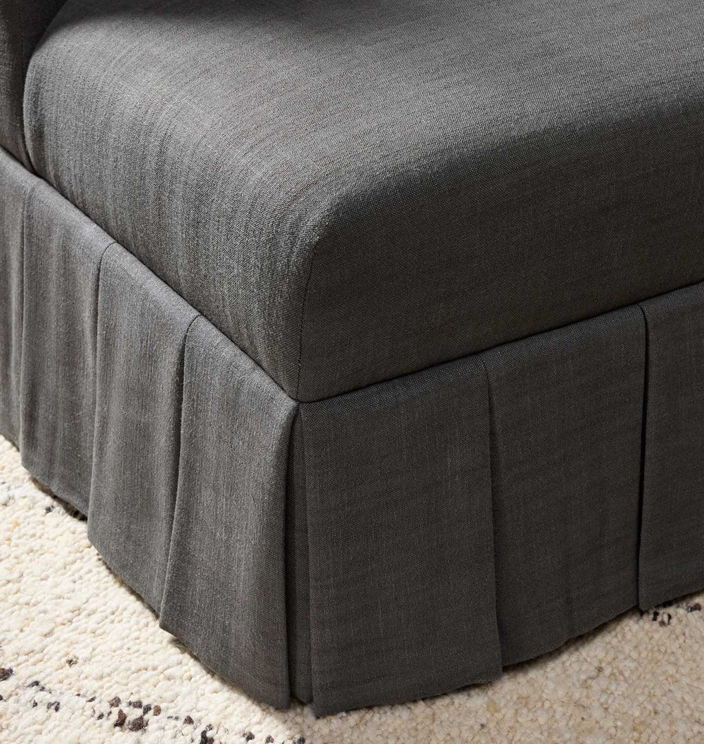 A close-up of a dark gray upholstered chair with a pleated skirt base, shown on a light beige textured rug.