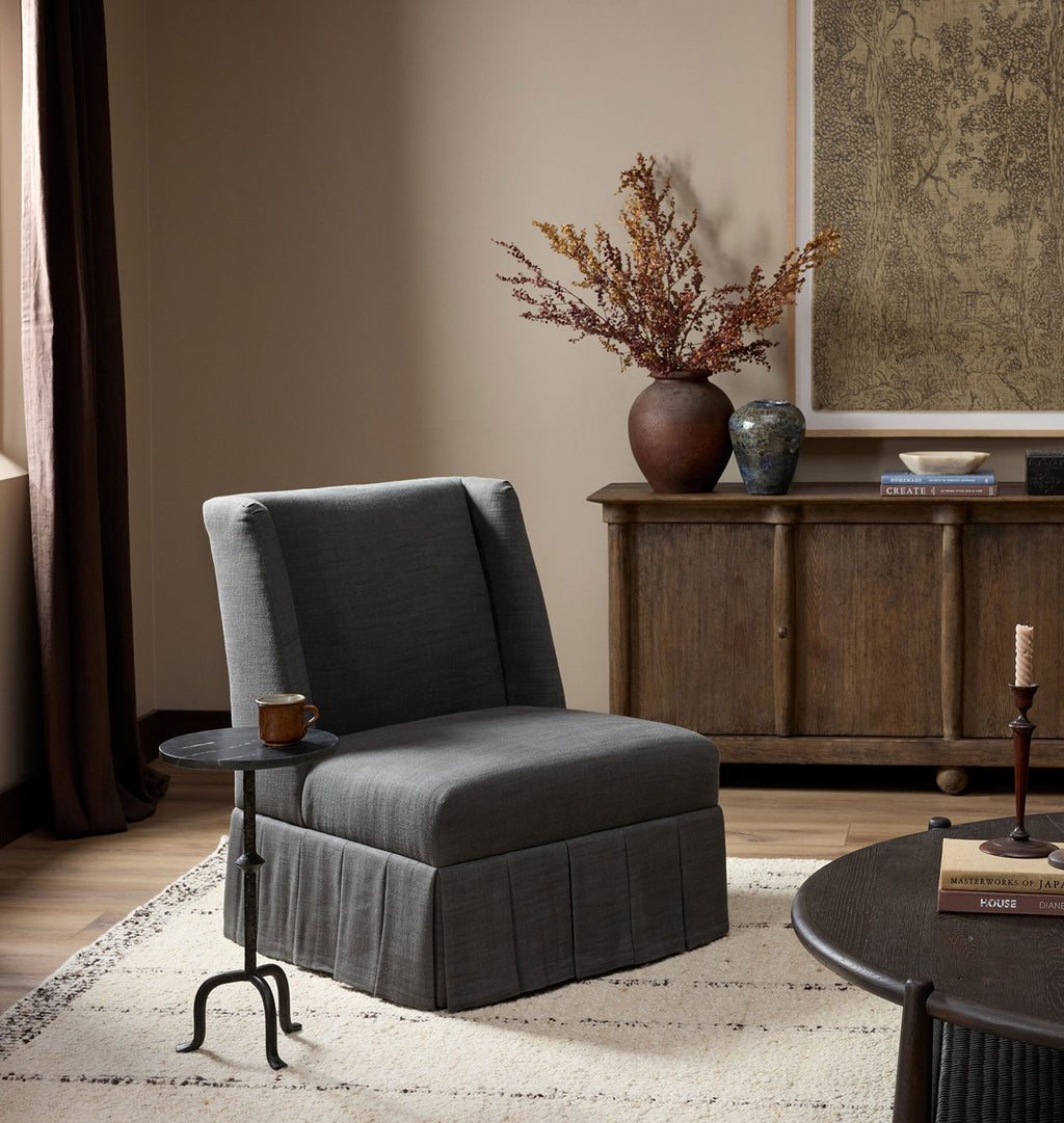 A dark gray upholstered armchair with a high back and skirted base, styled in a warm, modern living room setting next to a small side table.