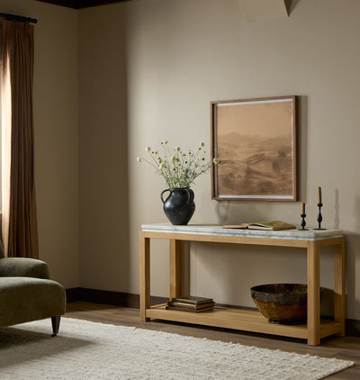 A light natural wood console table with a white marble top is styled in a warm, neutral-toned living room. The table features an open lower shelf holding books and a decorative bowl, and is styled with a black ceramic vase of wildflowers, an open book, and candlesticks.
