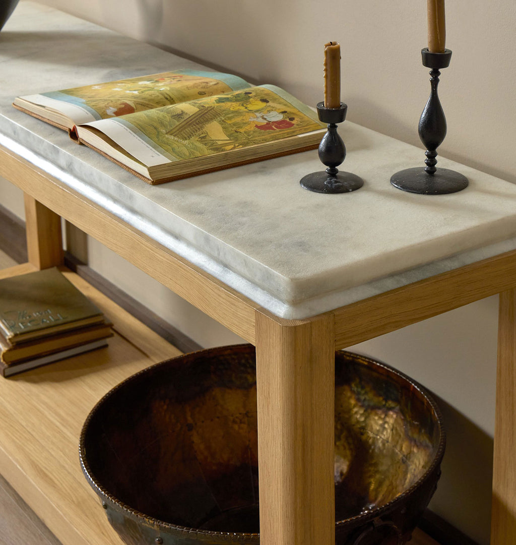 A wooden console table with a white marble top, styled with an open book, two black candlesticks, and a large hammered bronze bowl on the shelf below.