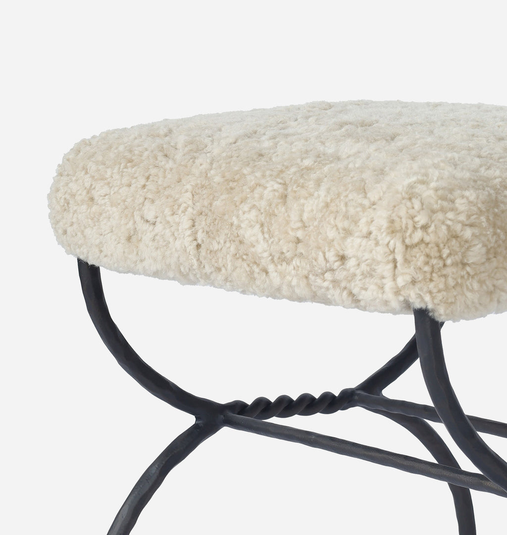 Close-up of a stool with a thick, fluffy cream-colored sheepskin seat cushion and a black metal frame, set against a plain white background.