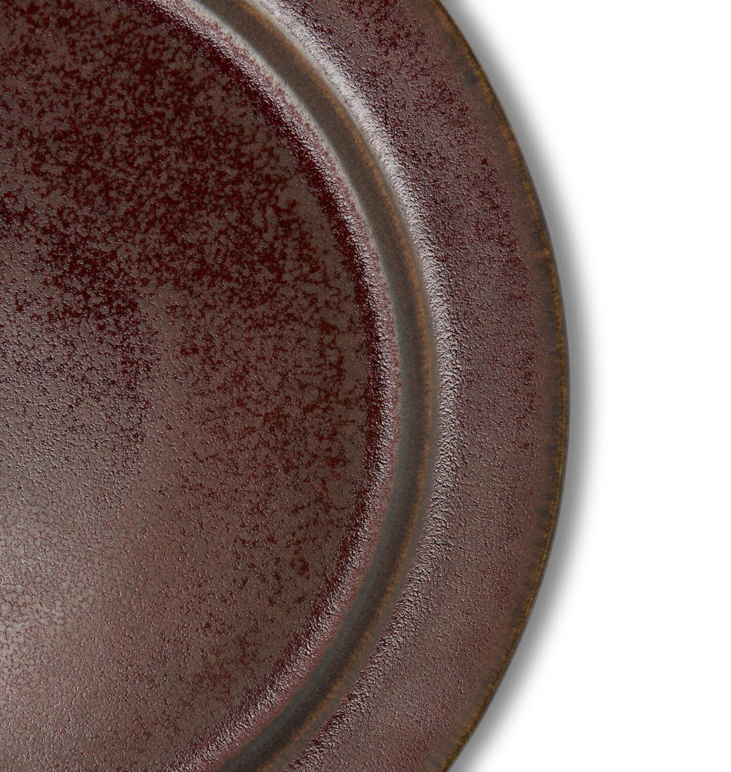 A close up detail shot of a round ceramic salad plate in a dark reddish brown glaze shown on a white background