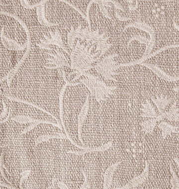 A close-up of a woven embroidered fabric in a warm taupe-beige color, featuring a floral and scroll pattern.