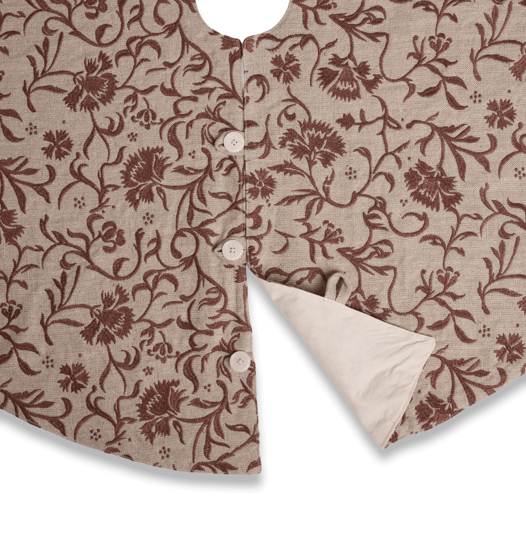 A close up detail of a round beige tree skirt with a reddish brown embroidered floral and scroll motif pattern with three button closures set against a white background
