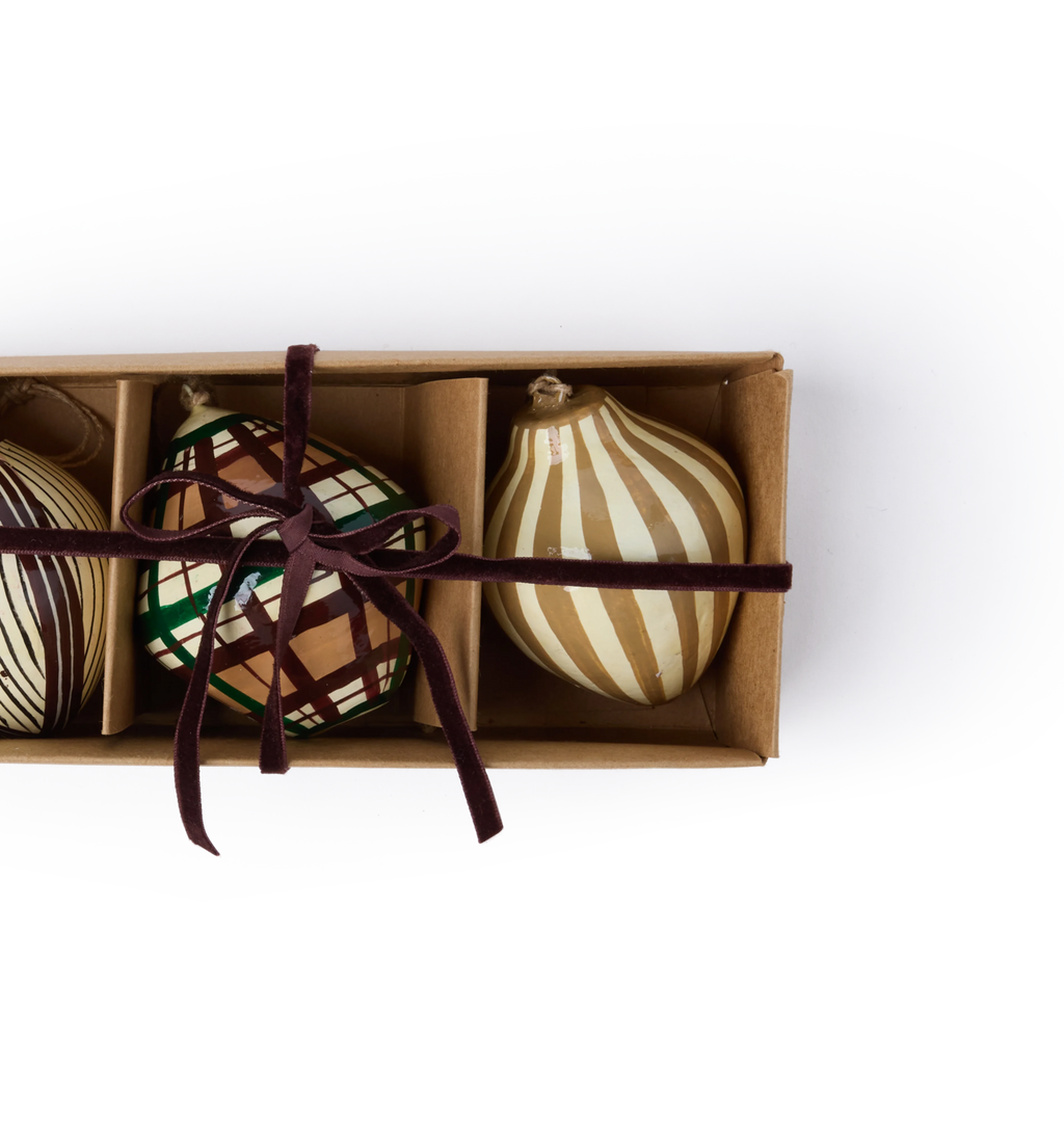 A set of decorative paper mache ornaments with striped and plaid patterns and red velvet ribbon in a gift box