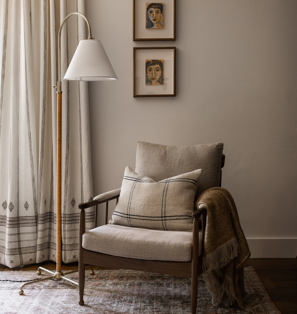 A styled mid-century modern armchair in a warm neutral room, featuring a beige linen seat and back cushion, a plaid throw pillow, a brown fringed throw blanket, a tall floor lamp, and patterned curtains.