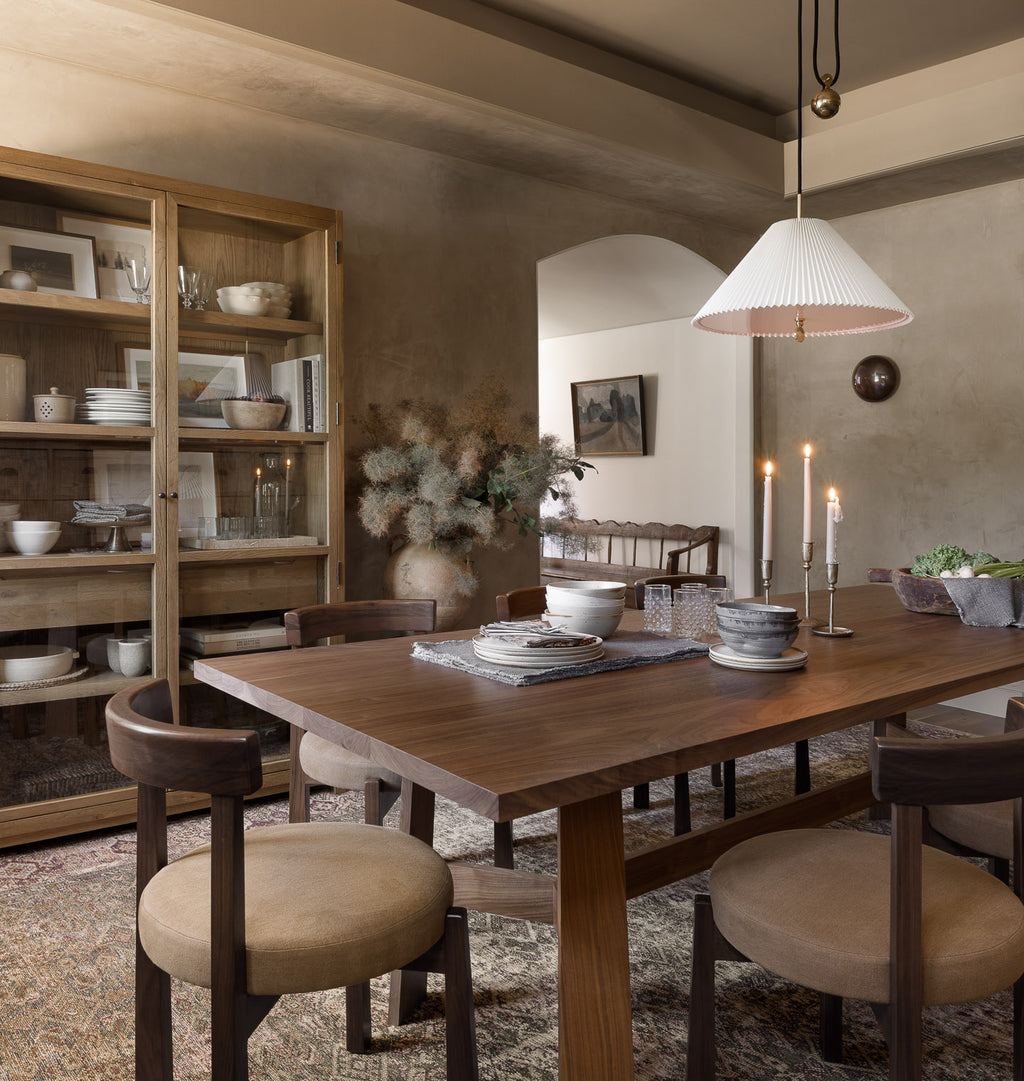 A rustic wooden dining table with upholstered chairs in a warm, neutral-toned dining room. A white ribbed pendant light hangs above, and a large wooden hutch filled with dishes stands to the left.