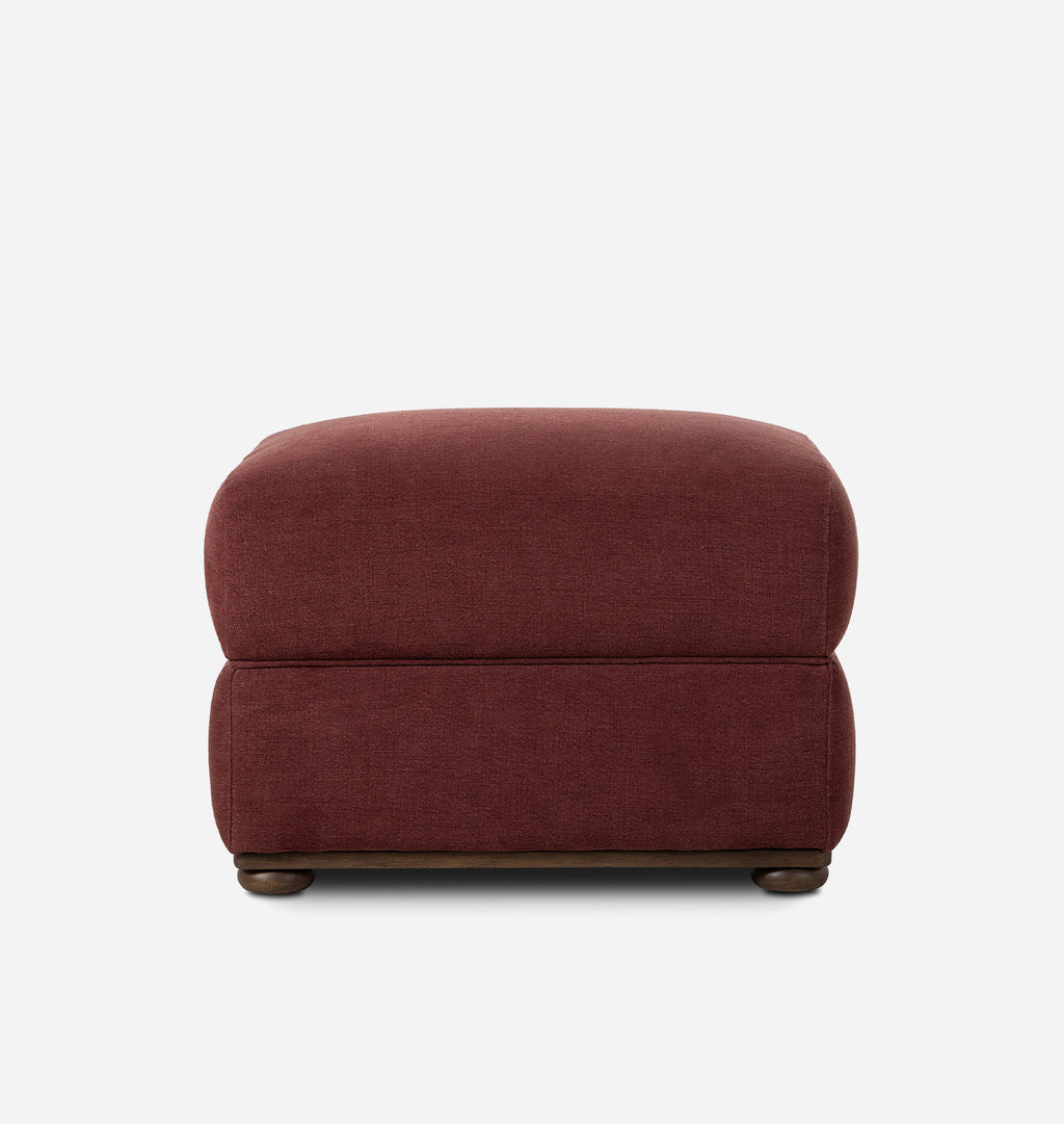 A square upholstered ottoman in a deep burgundy fabric, featuring dark wood feet, shown against a plain white background.