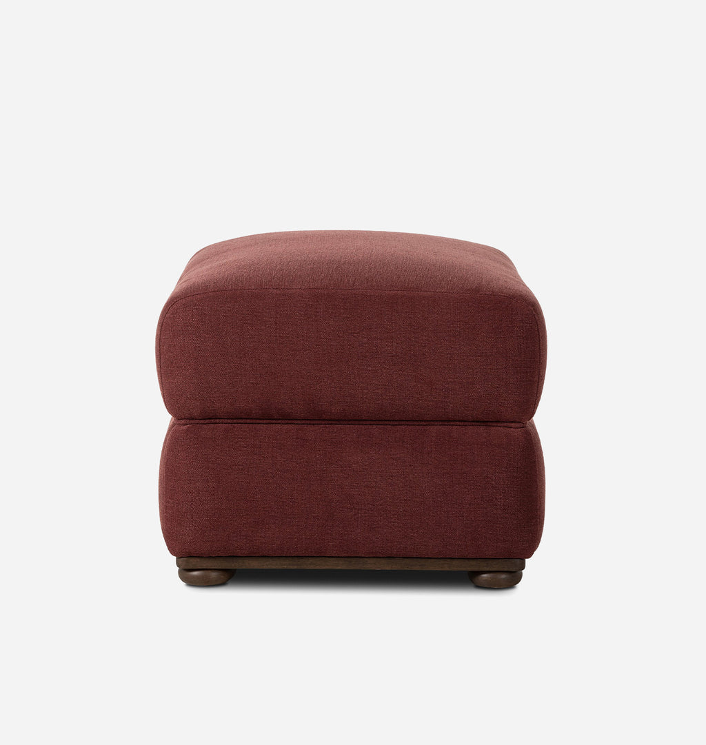 A square upholstered ottoman in a textured rust-red fabric, featuring a dark wood base with rounded feet, shown against a plain white background.