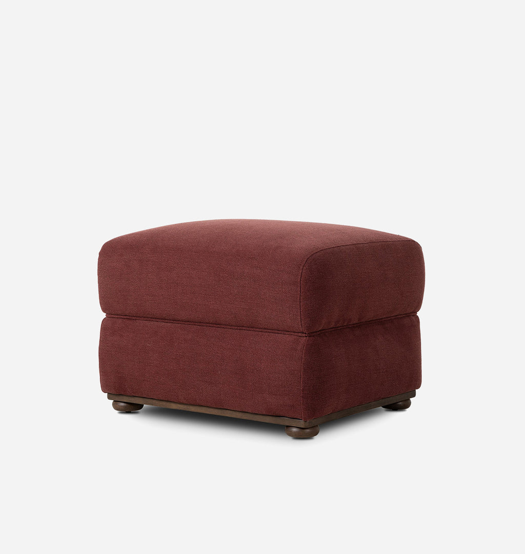 A square upholstered ottoman in a textured burgundy fabric, featuring dark wooden feet, shown against a plain light gray background.
