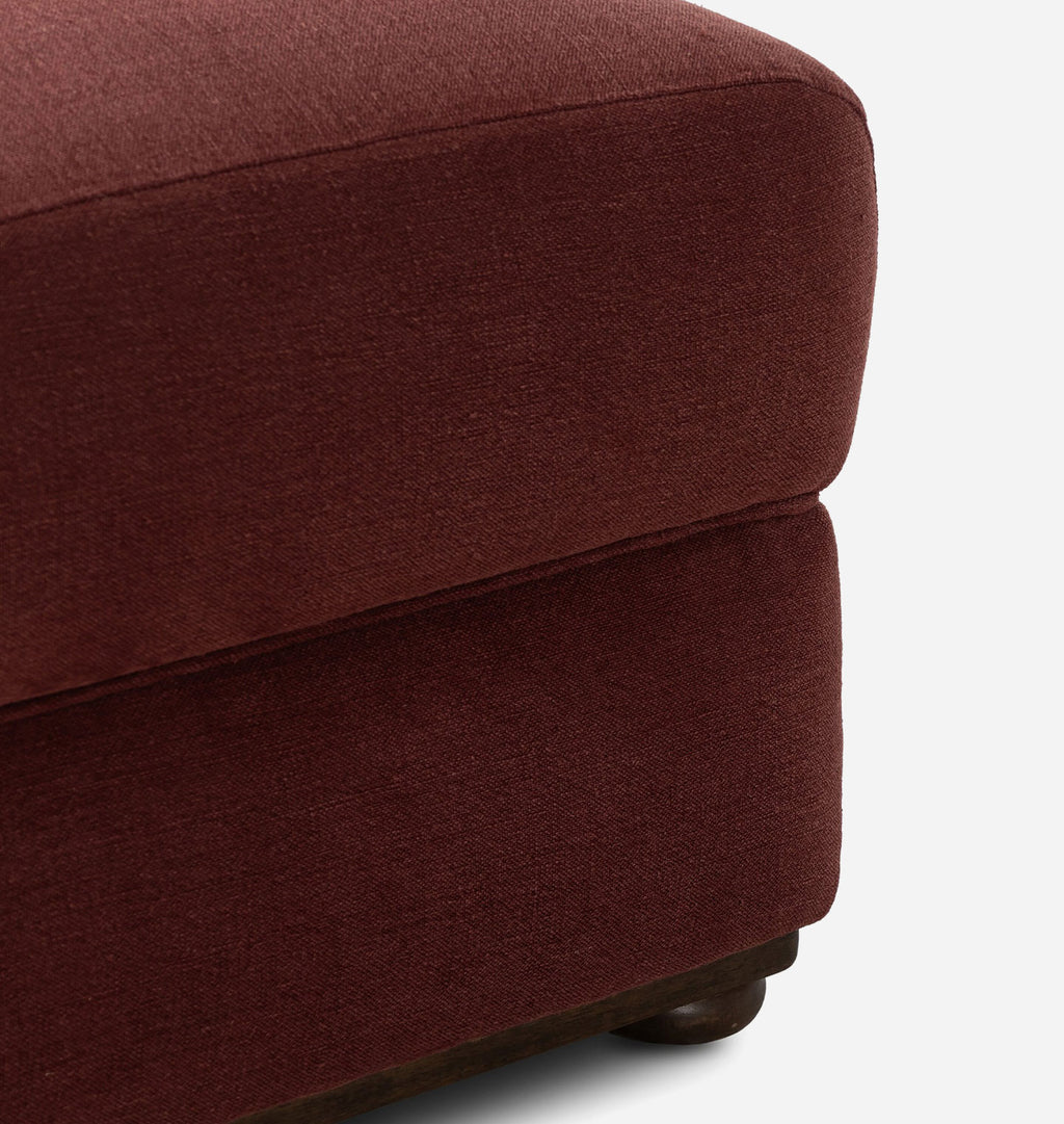 Close-up of a plush upholstered ottoman in a deep burgundy fabric, showing the textured weave and a small dark wooden foot visible at the bottom against a light gray background.