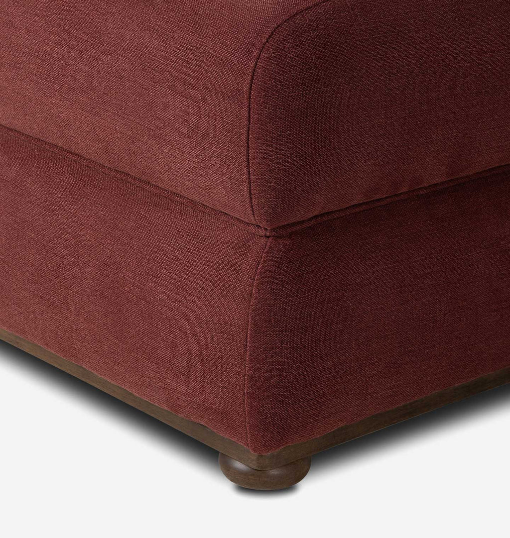 Close-up of the corner of a upholstered ottoman or sofa seat in a textured burgundy fabric, showing the corner seam, base, and a dark wood footstool leg on a white background.