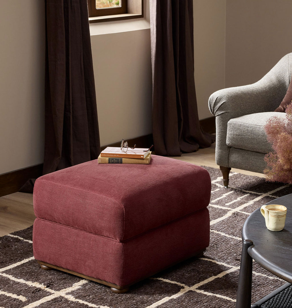 A square ottoman in textured dusty rose fabric, styled in a cozy living room setting with a gray armchair, dark curtains, a patterned rug, and a side table.