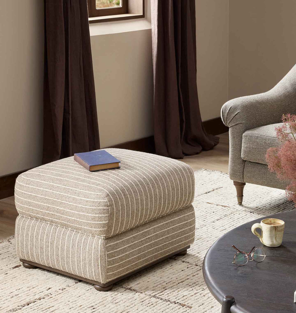 A square upholstered ottoman in beige fabric with thin vertical white stripes, styled in a living room setting with a blue book on top, next to a gray armchair and a dark coffee table.