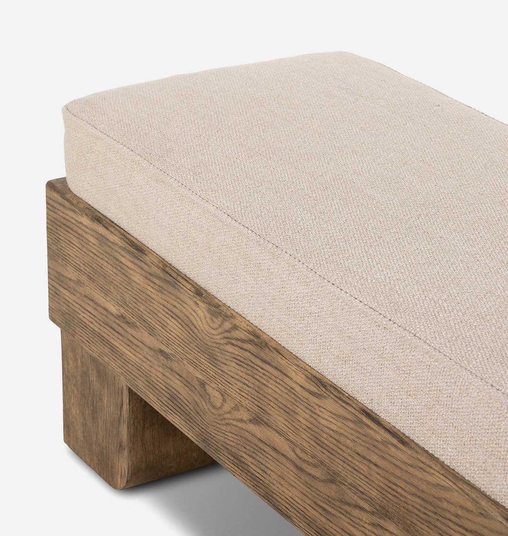 Close-up of a wooden bench or ottoman with a thick, textured beige fabric cushion, showing the wood grain and the corner of the seat against a clean white background.