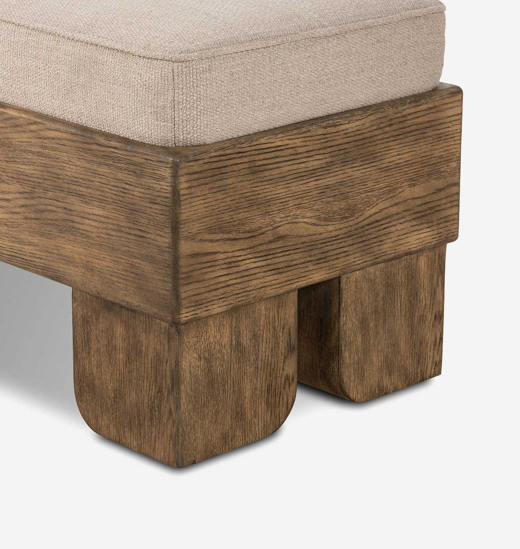 Close-up of an upholstered bench showing a light beige textured fabric seat cushion and a rustic medium-brown wooden frame with blocky legs, on a white background.