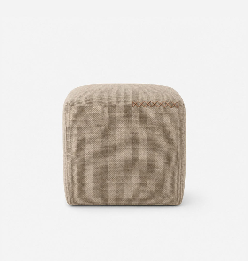 A square, low-profile pouf or ottoman upholstered in a beige textured fabric, featuring leather x-stitching on the top surface, set against a seamless white background.