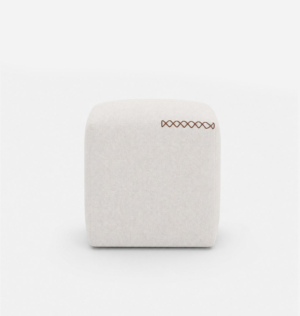 A square, low-profile pouf or ottoman upholstered in a light beige textured fabric, featuring leather x-stitching on the top surface, set against a seamless white background.