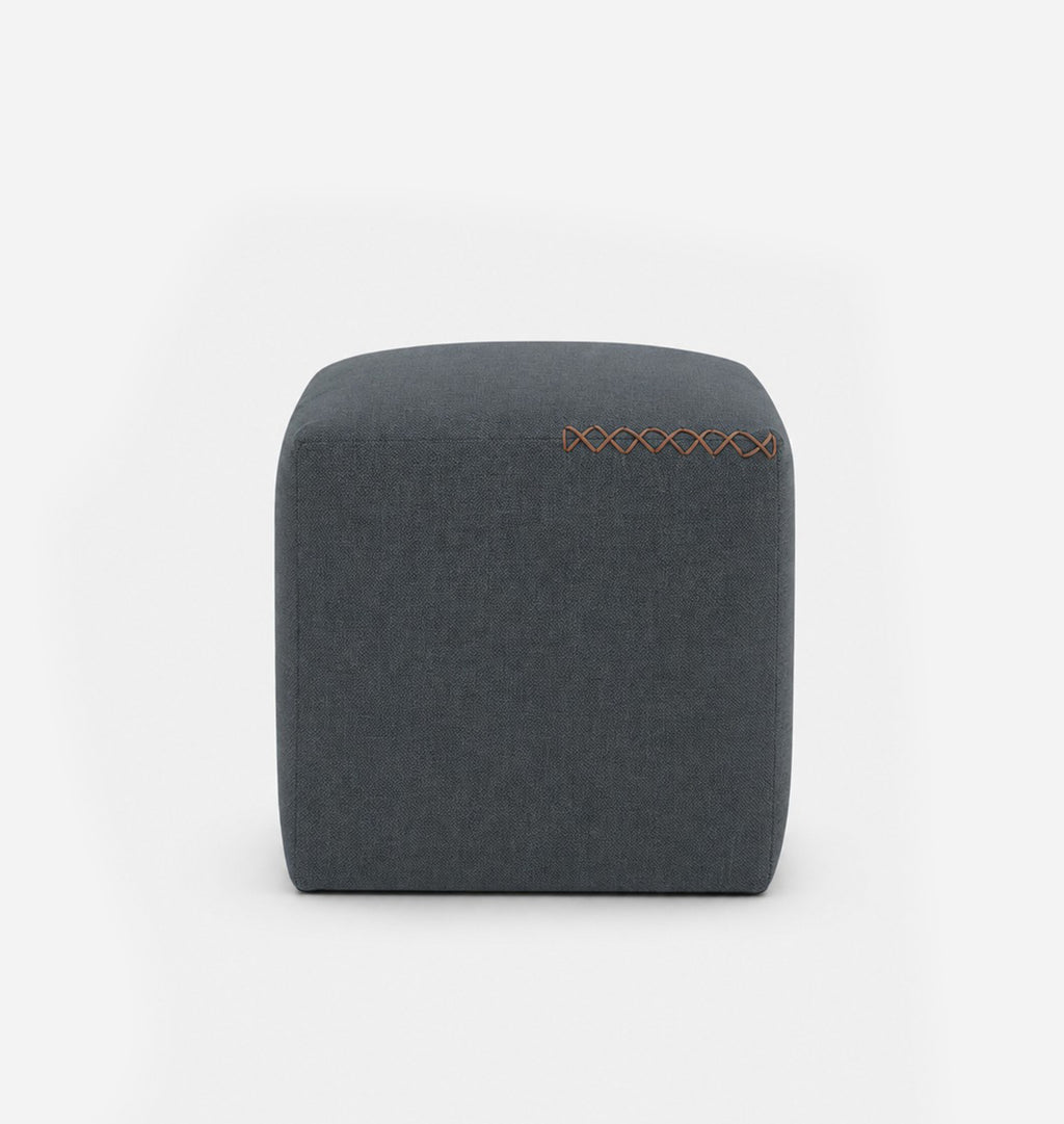 A square, low-profile pouf or ottoman upholstered in a dark muted blue grey textured fabric, featuring leather x-stitching on the top surface, set against a seamless white background.