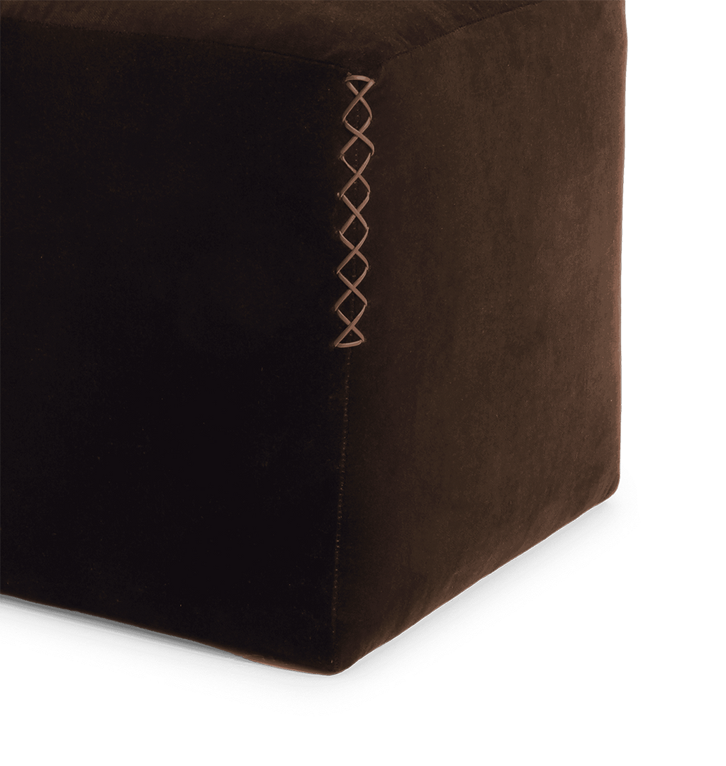 Close-up of a dark brown upholstered ottoman or cube stool corner, showing a x-stitched seam on a white background.
